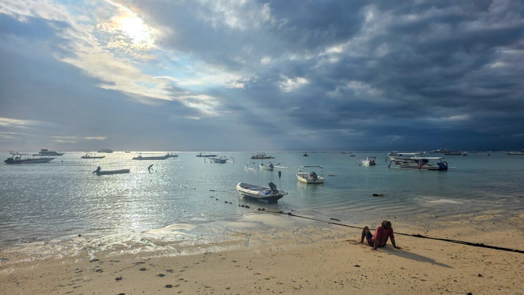 Man sitting on the beach in Nusa Lembongan looking toward the ocean as sunlight breaks through heavy clouds.