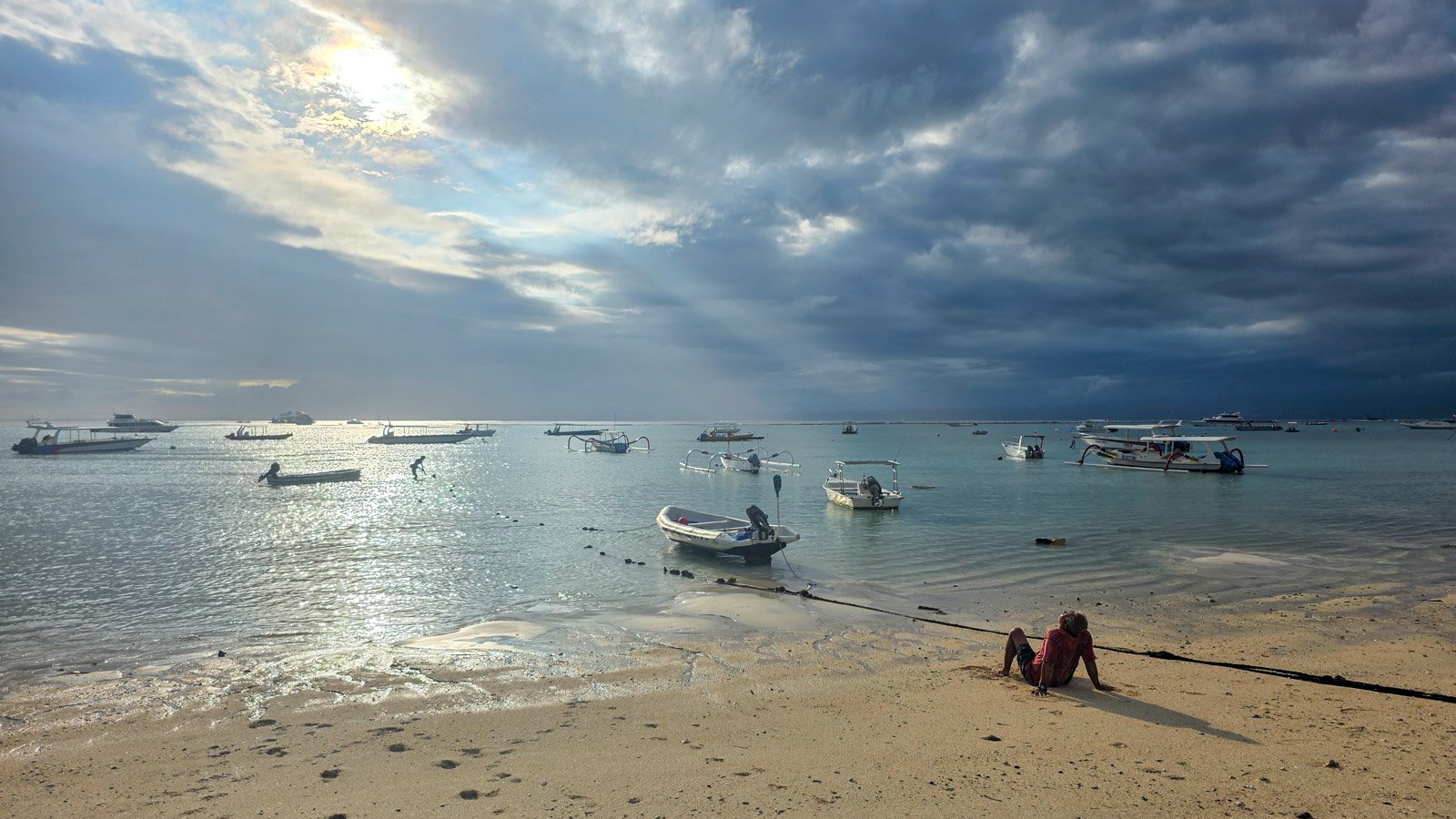 Man sitting on the beach in Nusa Lembongan looking toward the ocean as sunlight breaks through heavy clouds.