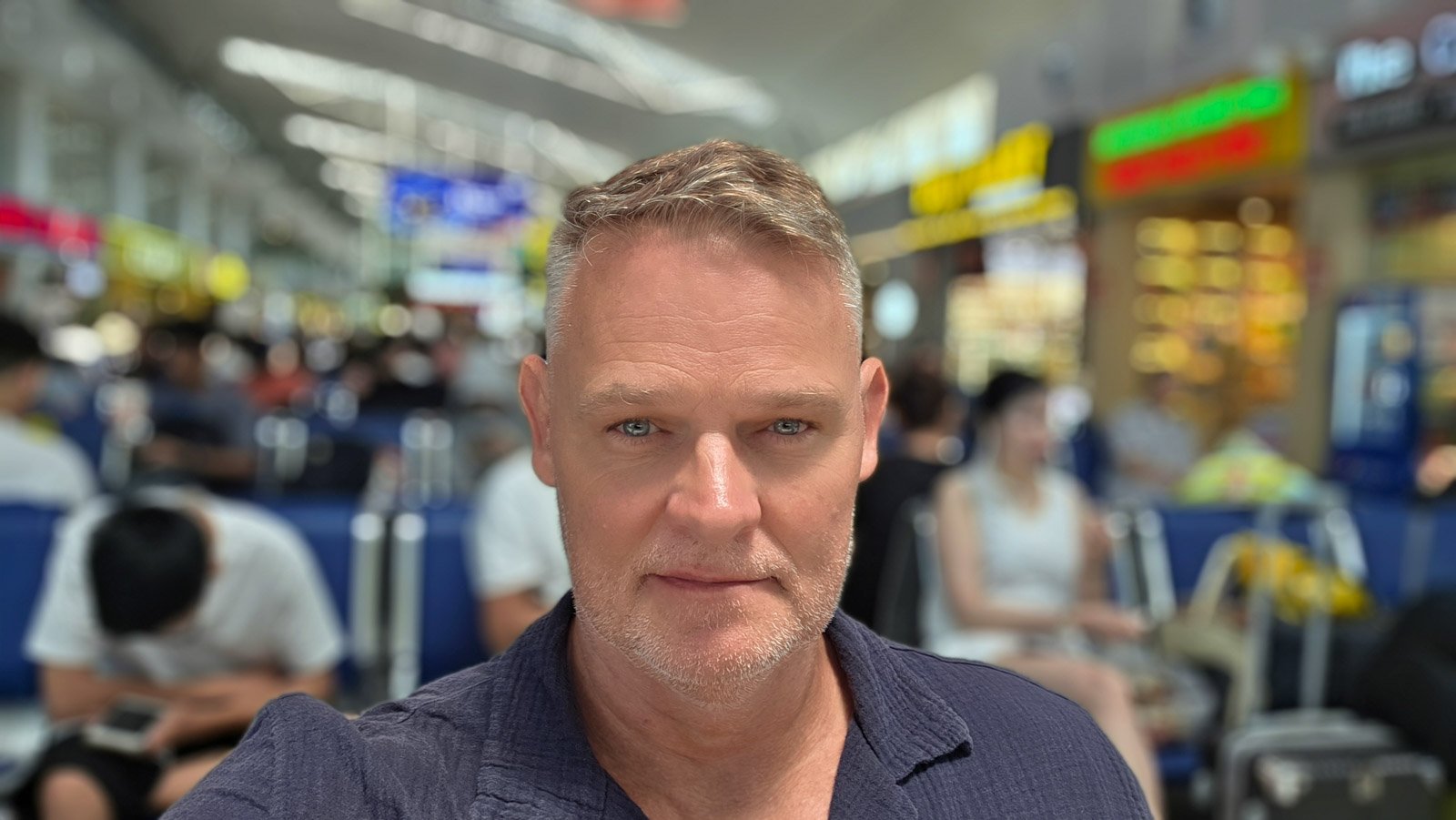Martin Gram sitting on a bench at Da Nang airport with travelers blurred in the background.