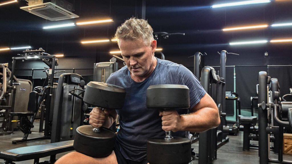 Man sitting on a gym bench holding heavy dumbbells and looking down, illustrating procrastination before taking action