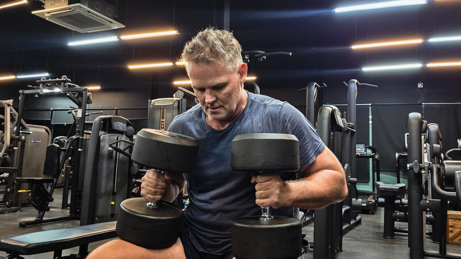Man sitting on a gym bench holding heavy dumbbells and looking down, illustrating procrastination before taking action